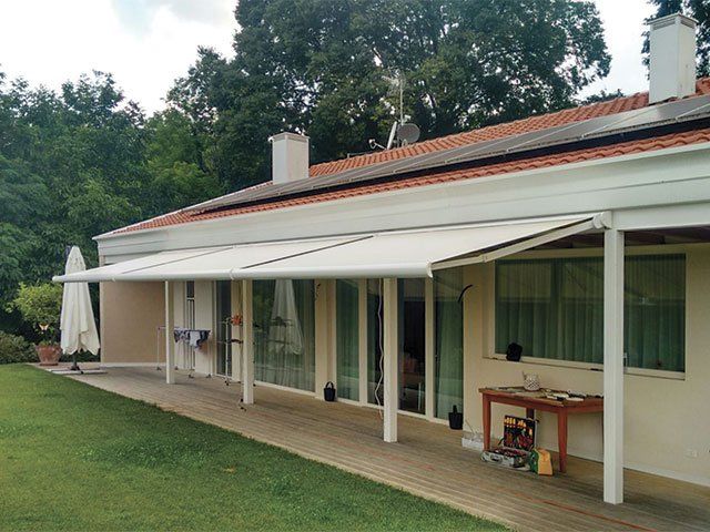 Una tenda bianca si estende su un patio con finestre, proteggendo dalla luce solare. Prato verde, alberi e casa color crema.