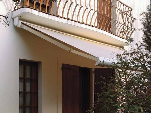 Tenda da sole bianca che si estende da un balcone e copre una porta. Persiane marroni e ringhiera in ferro battuto.