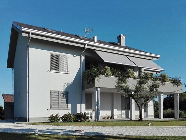Casa bianca a due piani con balcone, tende da sole e un piccolo albero di fronte, che si staglia contro un cielo azzurro.