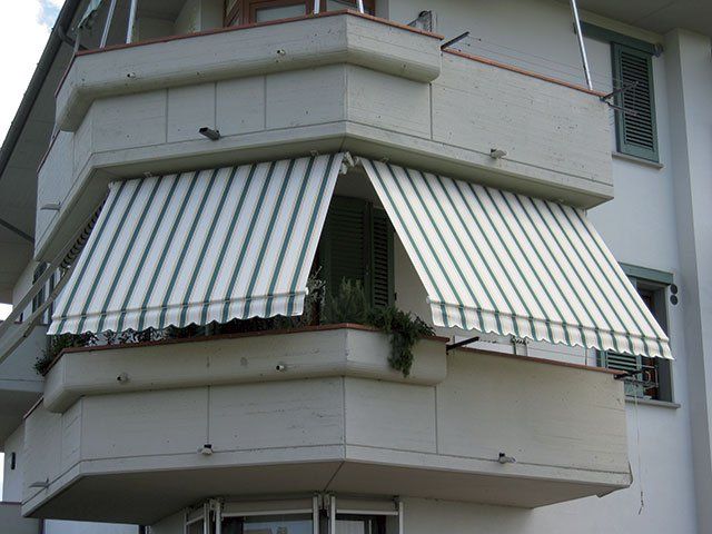 Una tenda a strisce bianche e verdi copre un balcone angolare di un edificio di colore chiaro.