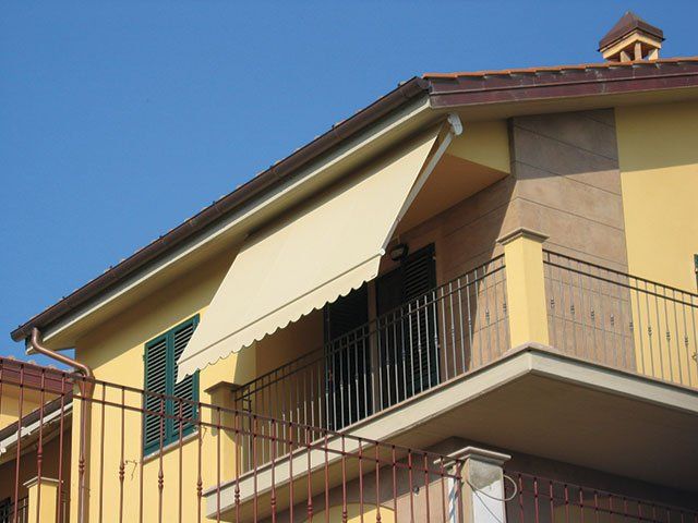 Una tenda beige si estendeva su un balcone con ringhiera nera, sullo sfondo di un edificio giallo e di un cielo azzurro.