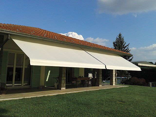 Tende da sole beige retrattili su un portico, prato verde, cielo azzurro.