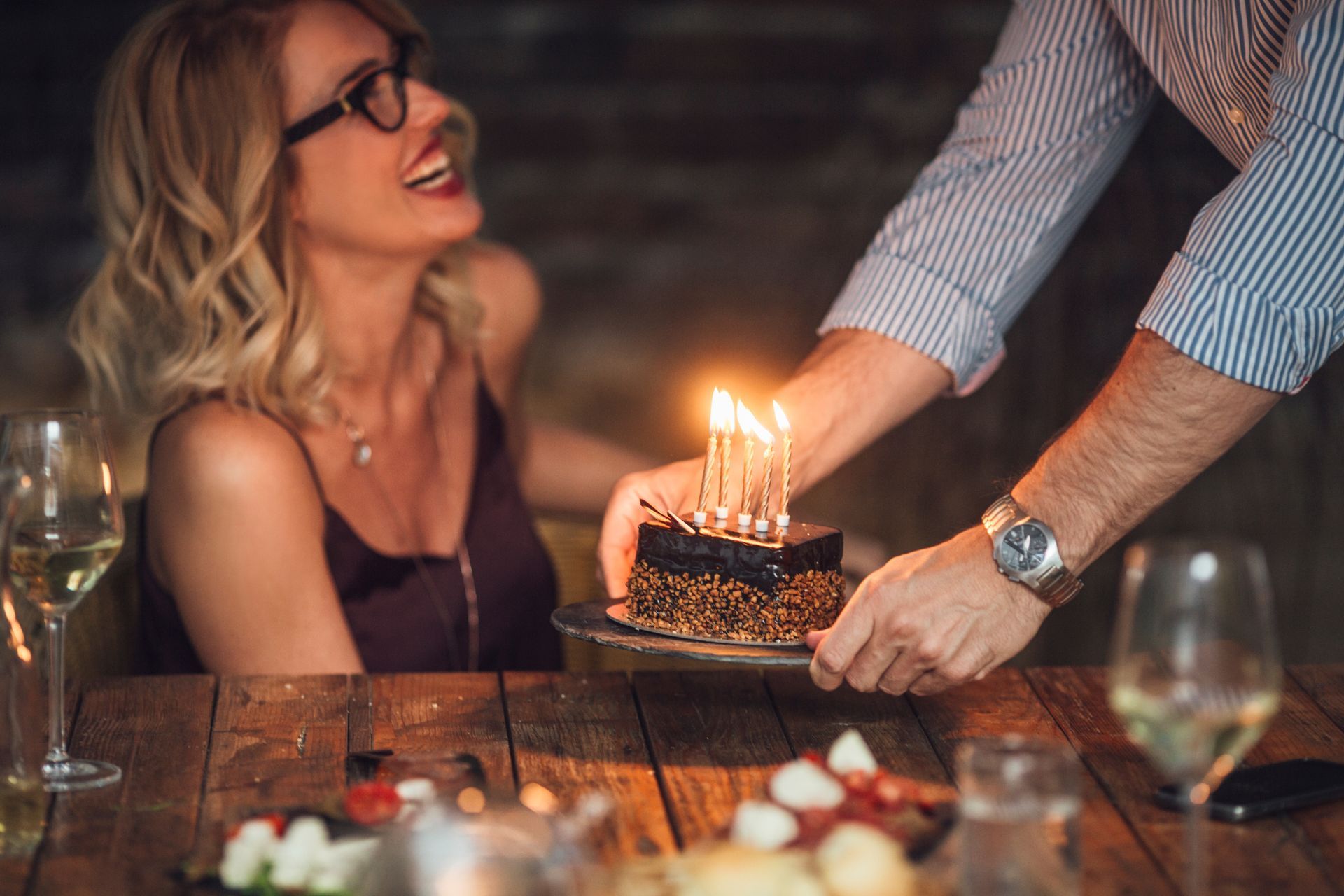 Birthday cake being presented at a candlelit dinner, with a smiling woman watching across the table.