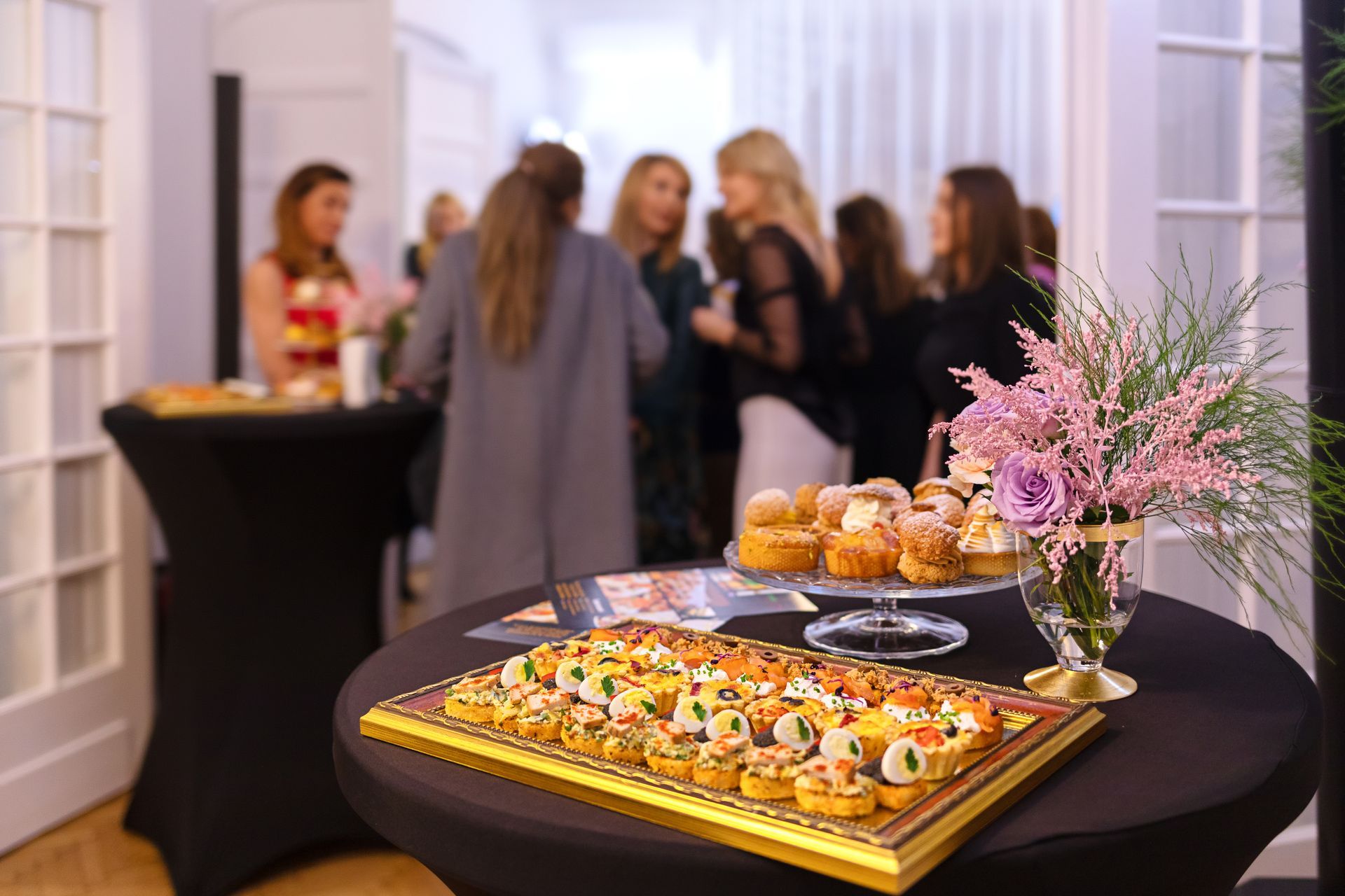Dessert table with pastries and pink flowers at a cocktail party, guests mingling in the background