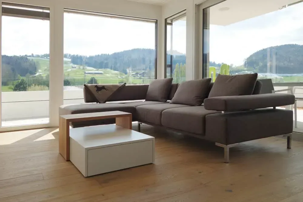 Modernes Wohnzimmer mit dunklem Ecksofa, niedrigen Couchtischen und großen Fenstern mit Blick auf grüne Hügel.