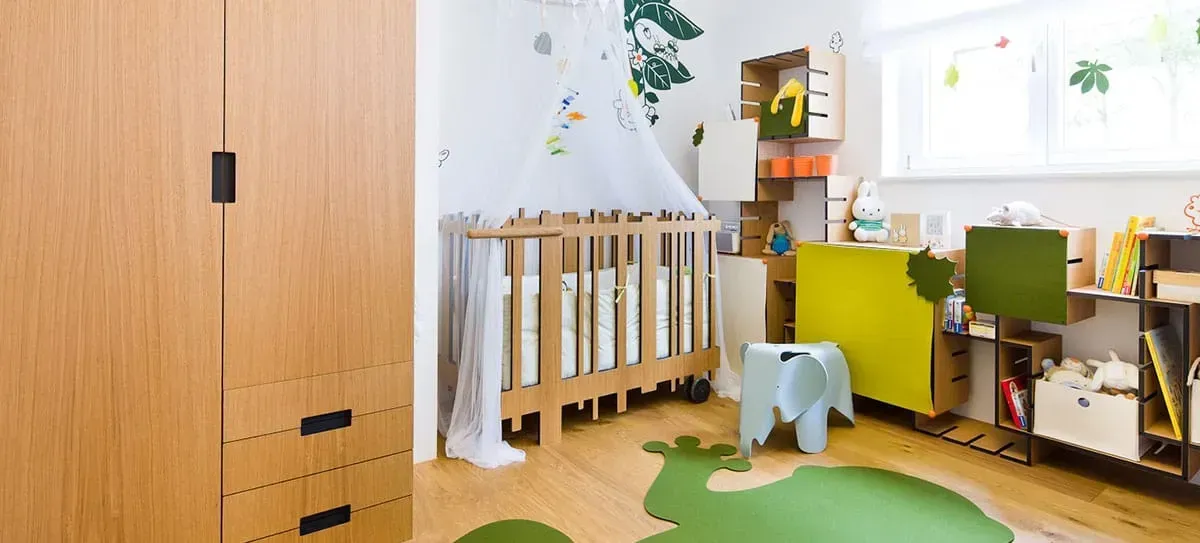 Heller Kita-Raum mit Holzkinderbett, Regalen und kleinem blauen Hocker auf einem grünen Teppich.