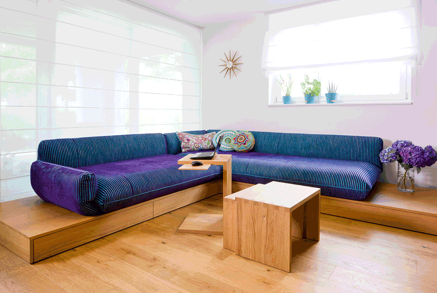 Lila Ecksofa in einem hellen Wohnzimmer mit Holztischen und Topfblumen