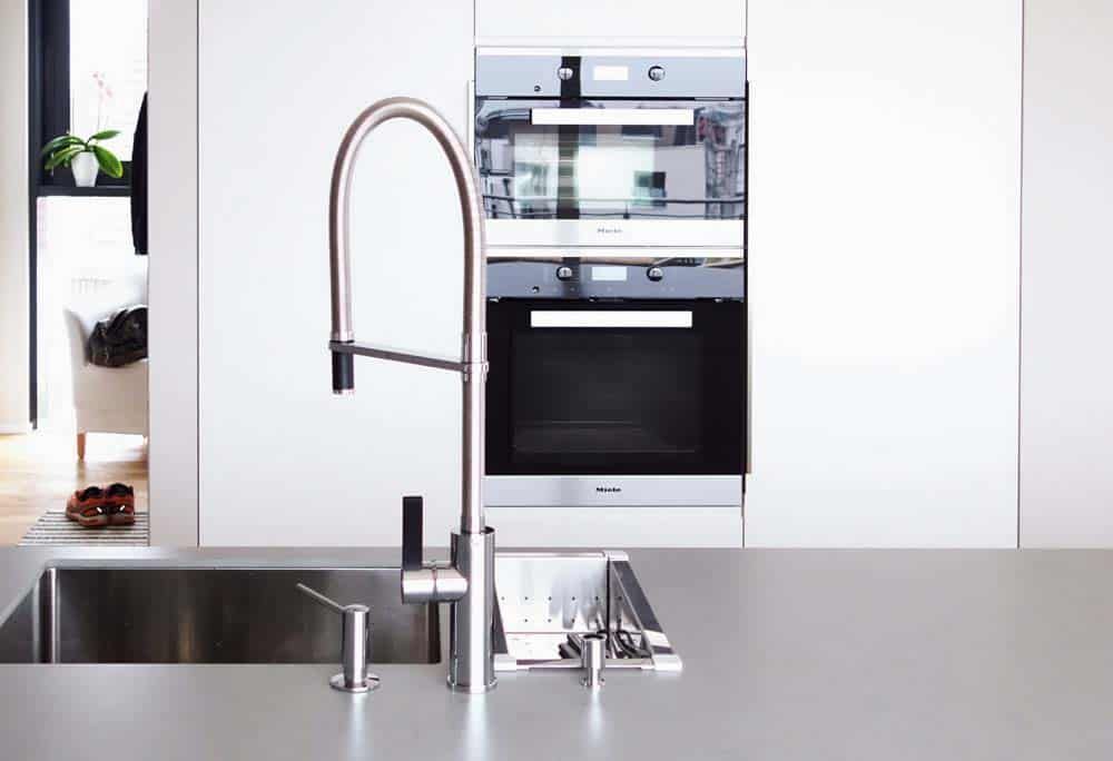 Moderne Küche mit Edelstahlspüle, hohem Wasserhahn und eingebautem Backofen hinter weißen Schränken.