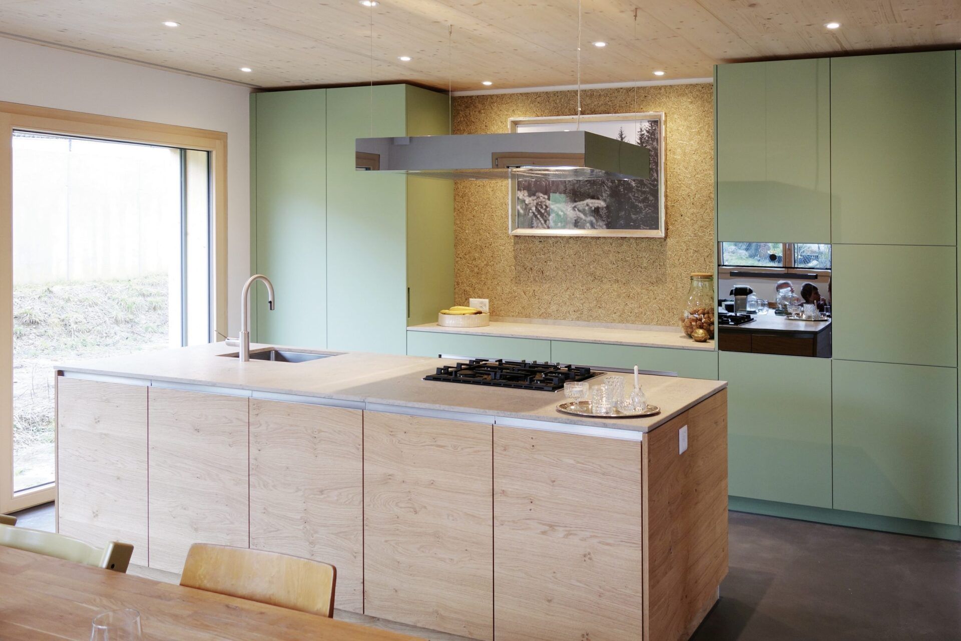 Moderne Küche mit salbeigrünen Fronten, Kücheninsel aus hellem Holz und integrierter Kochfläche, geplant und umgesetzt von Hase & Kramer Möbelwerkstätte GmbH in Dornbirn.