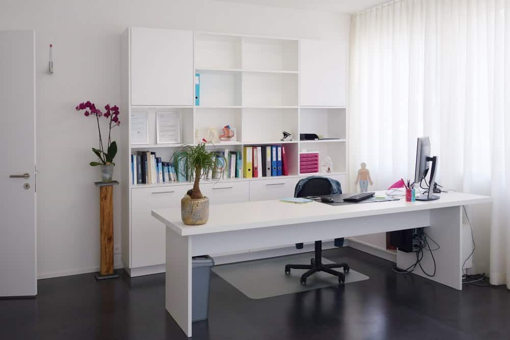 Modernes weißes Büro mit Schreibtisch, Regalen, Laptop, Büchern und Blumen an einem hellen Fenster