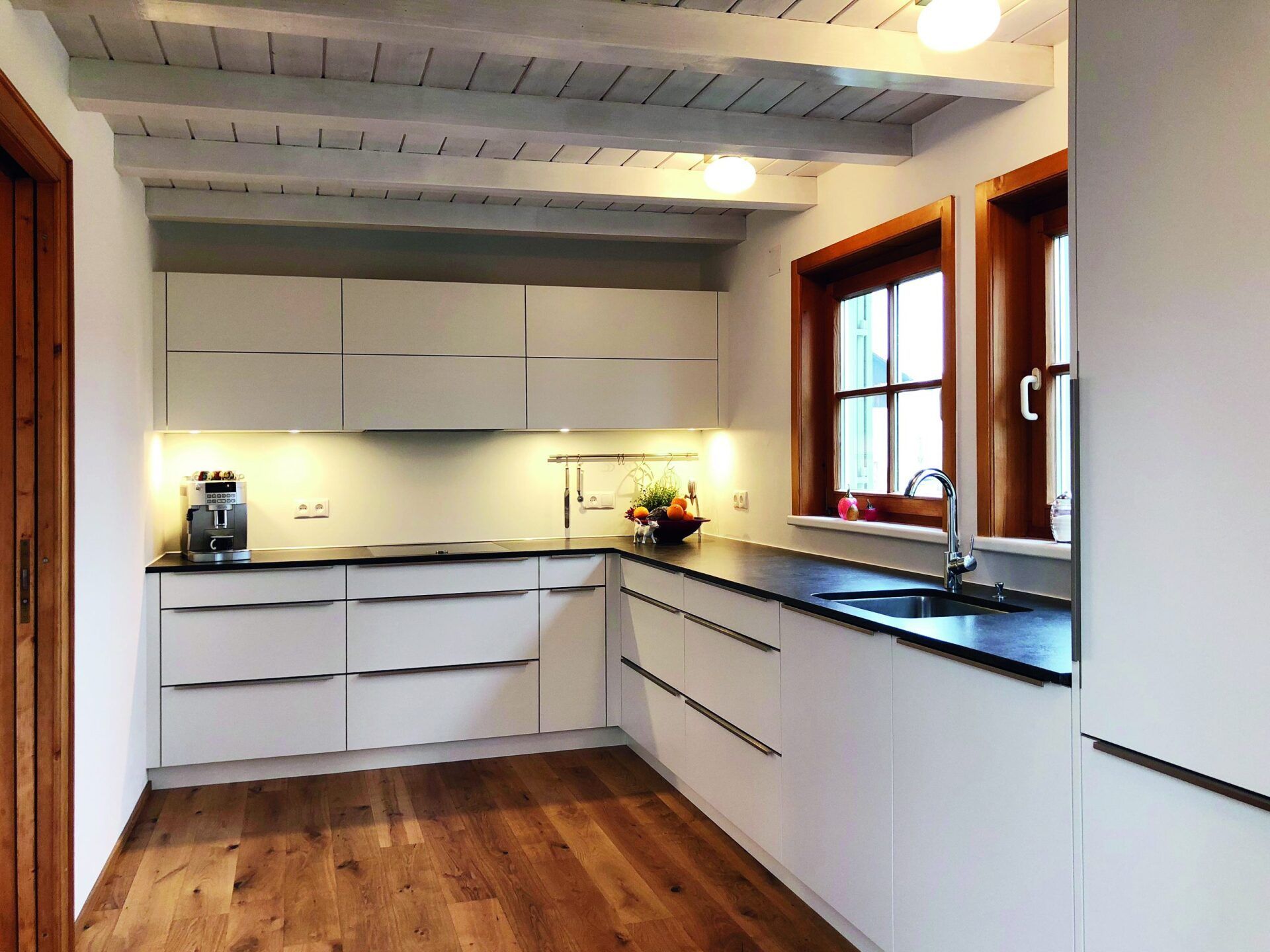 Helle, moderne Küche mit weißen Schränken, Holzboden, dunklen Arbeitsplatten und einem Fenster über der Spüle