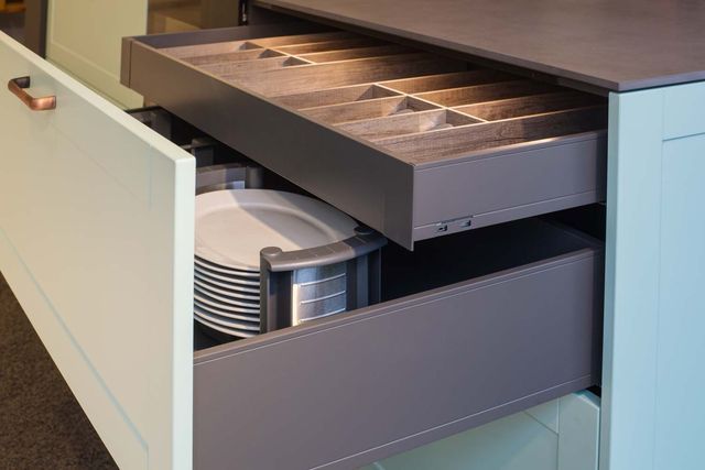 Offene Küchenschublade mit gestapelten Tellern und einem silbernen Besteckkasten in einem dunklen Schrank.