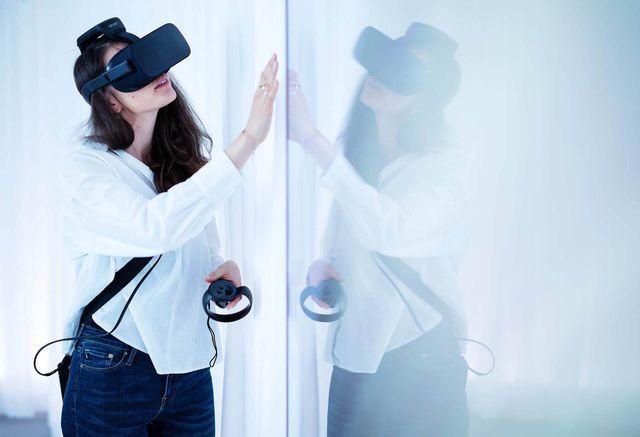Eine Frau mit VR-Headset greift nach einer Glaswand, in der ihr Spiegelbild sichtbar ist.