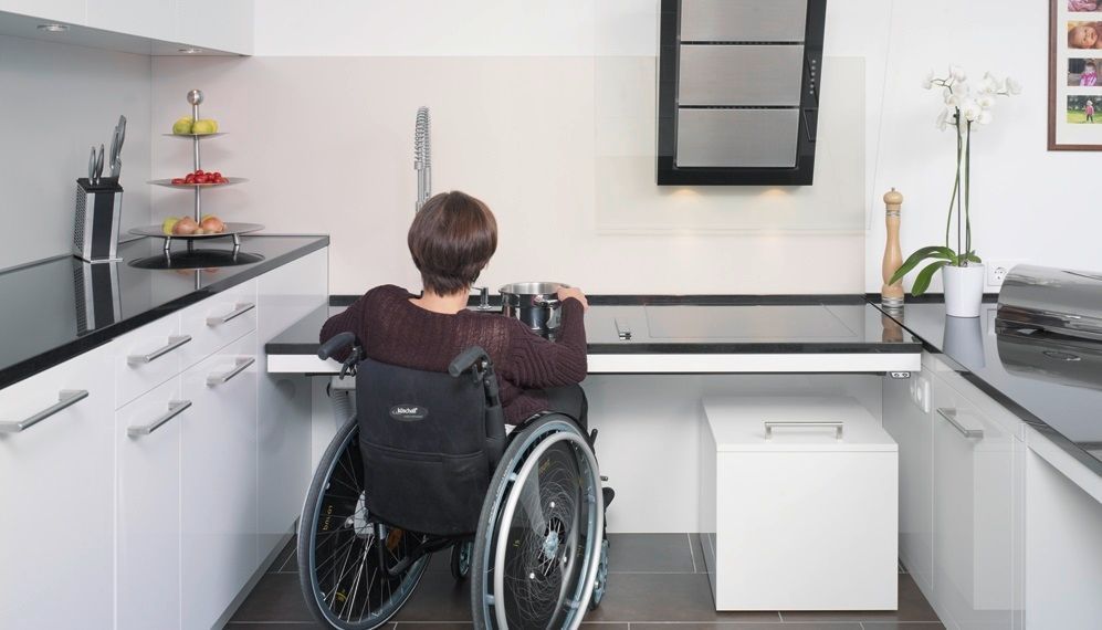 Eine Person im Rollstuhl steht an einer modernen Küchentheke und greift nach der Spüle.