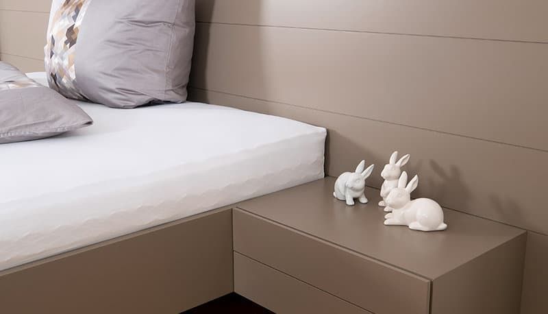 Minimalistisches Schlafzimmer mit weißer Bettwäsche und kleinen weißen Hasenfiguren auf einem taupefarbenen Nachttisch.