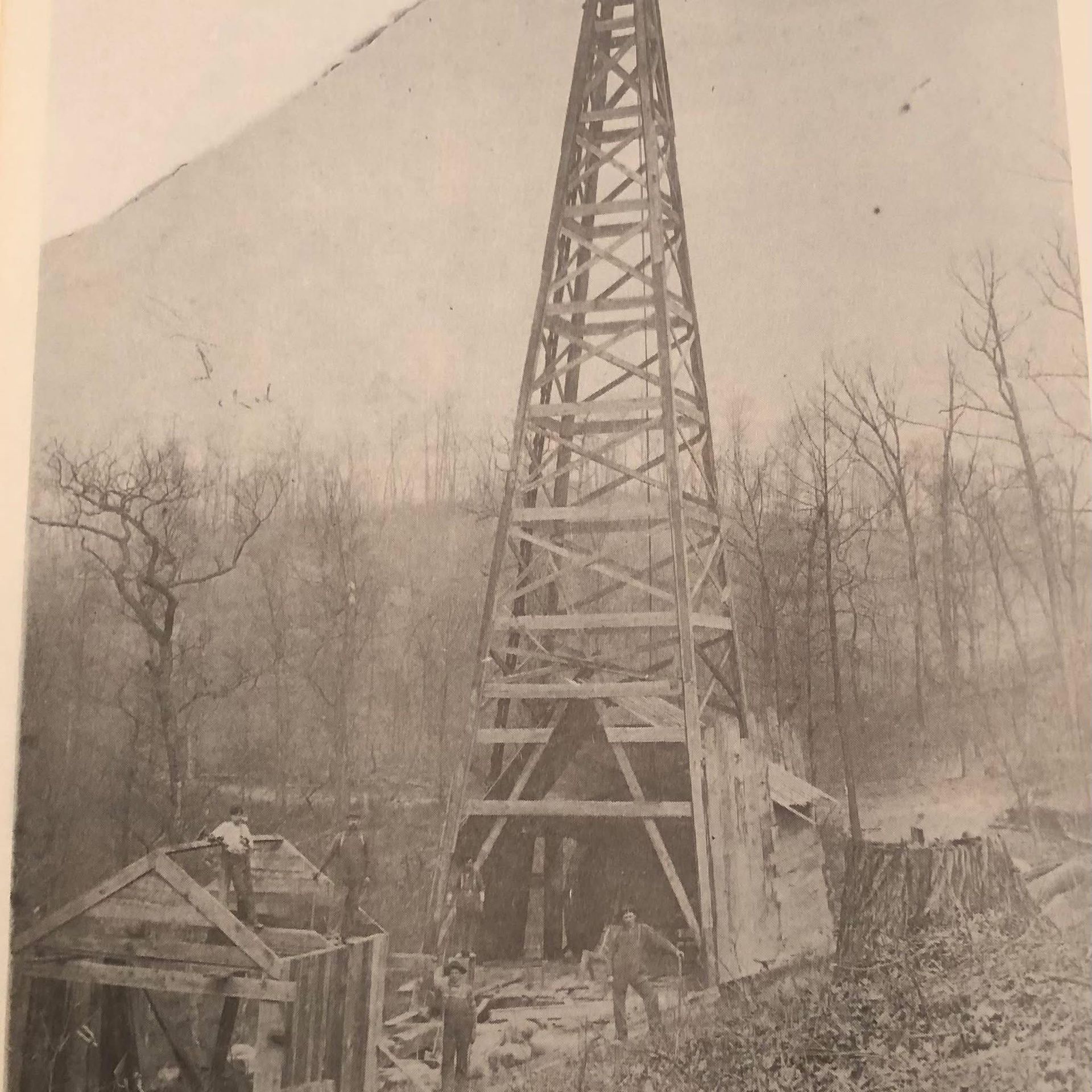 Wooden oil derrick in a wooded area, with several workers and a small building in front.