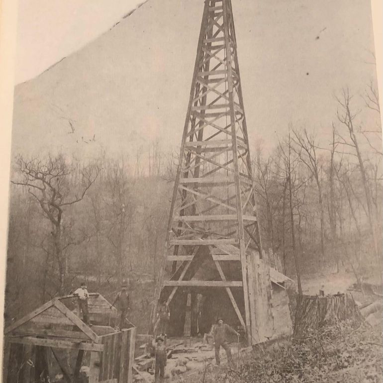 Wooden oil derrick in a wooded area, with several workers and a small building in front.