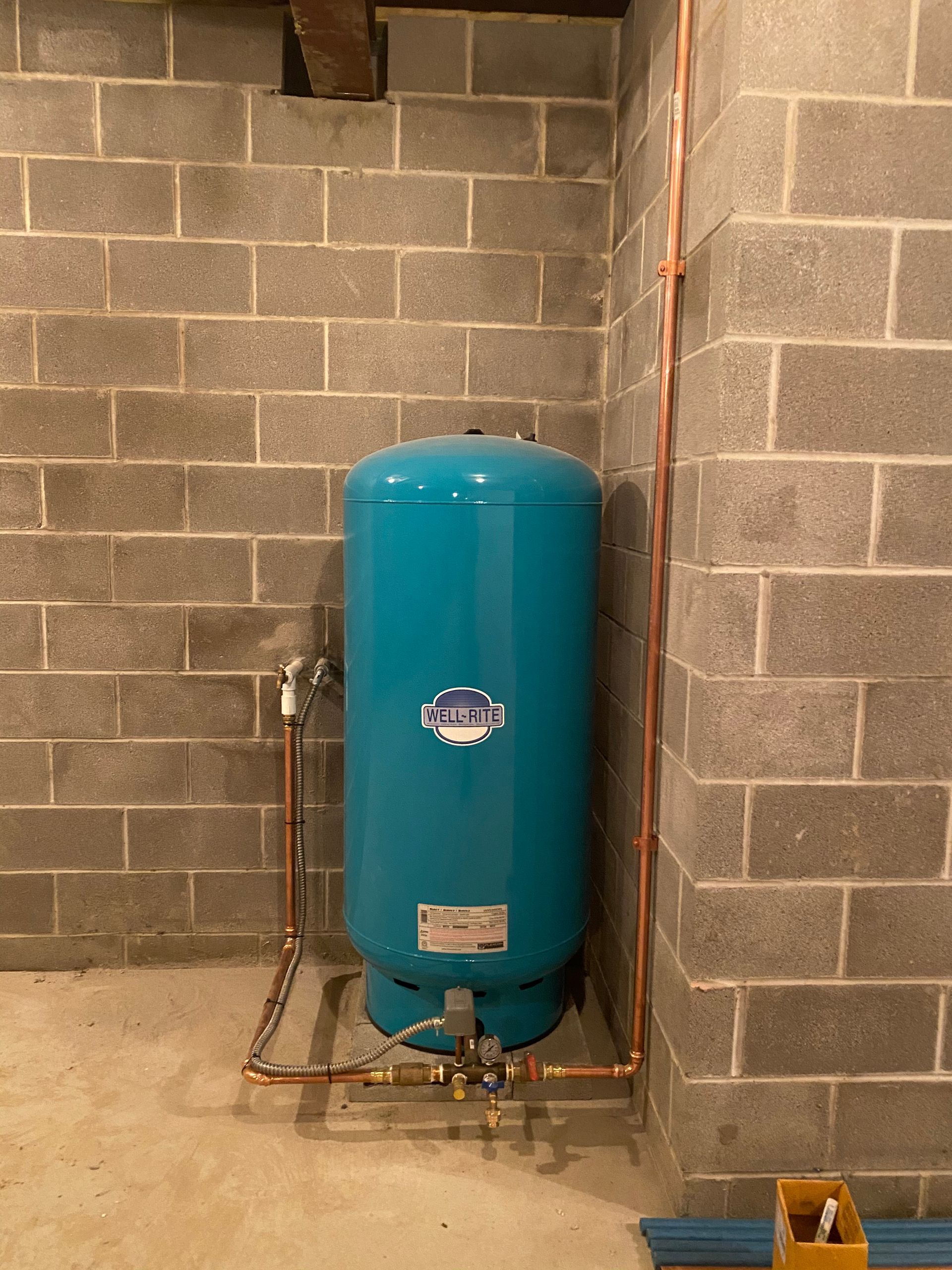 Blue water pressure tank in a basement corner, connected to copper pipes against a gray cinder block wall.