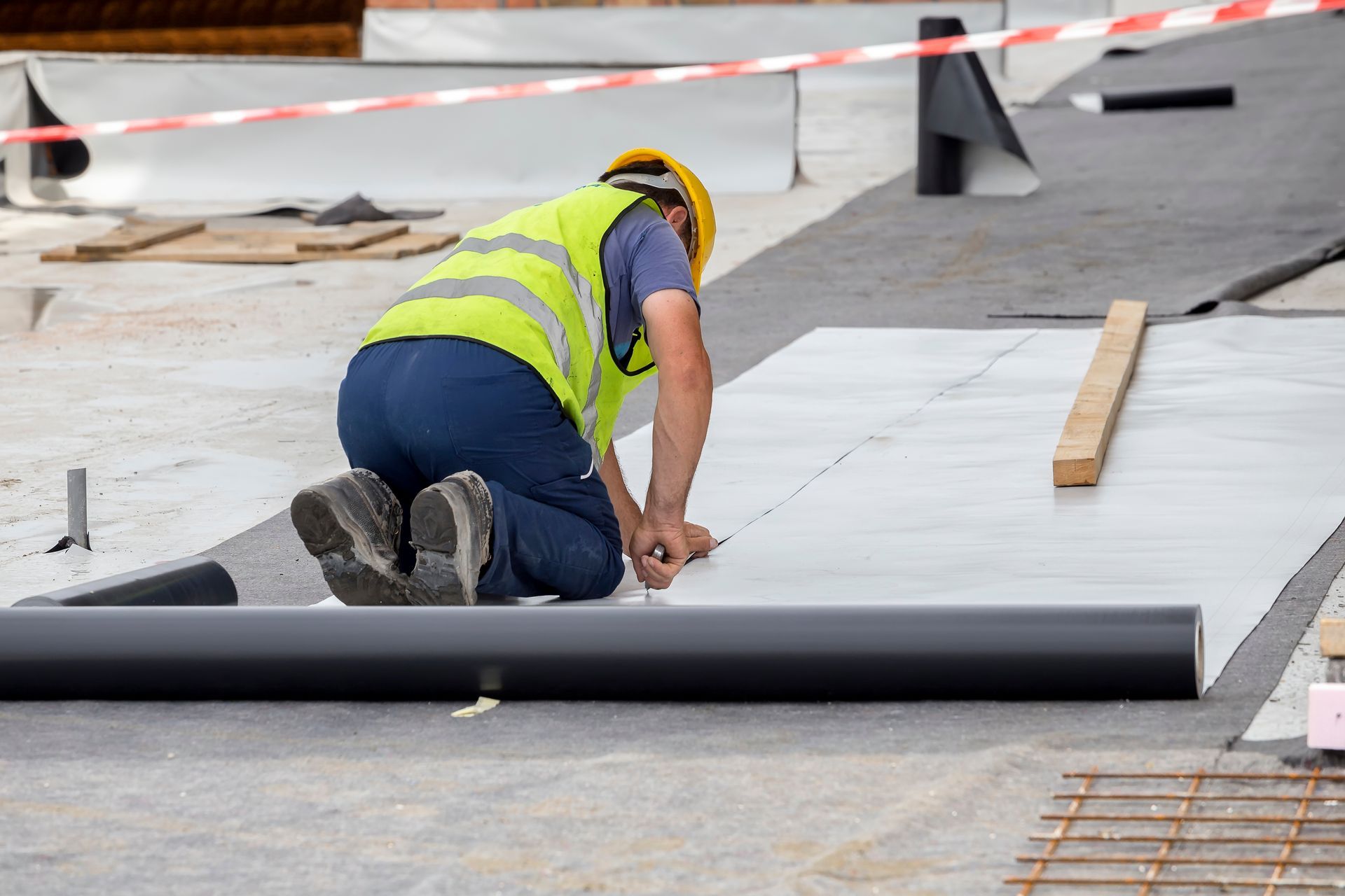 Un ouvrier du bâtiment, agenouillé, découpe des matériaux sur un toit plat, portant un gilet de sécurité