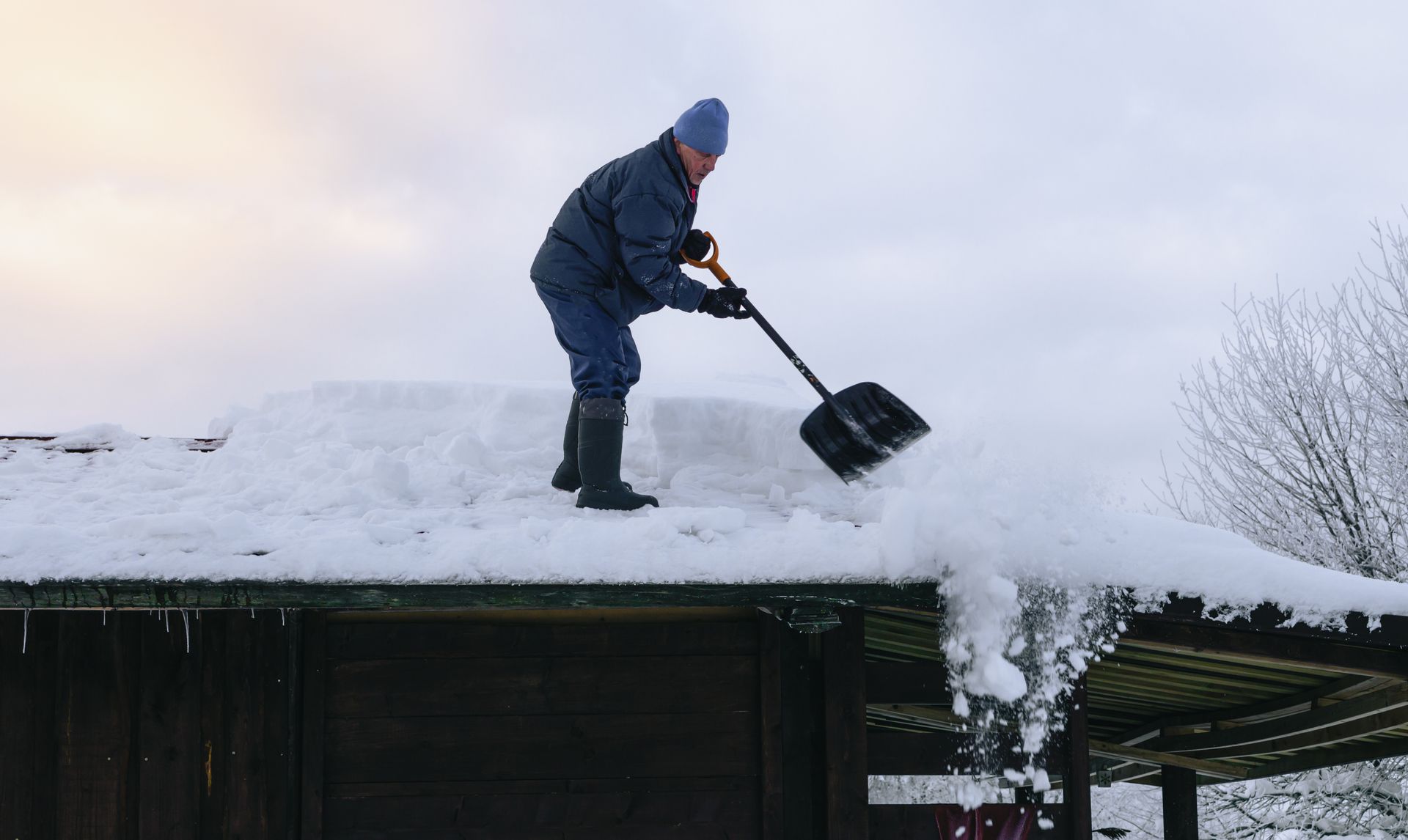Une personne déneige un toit, portant un chapeau et une veste bleus, sous un ciel nuageux.