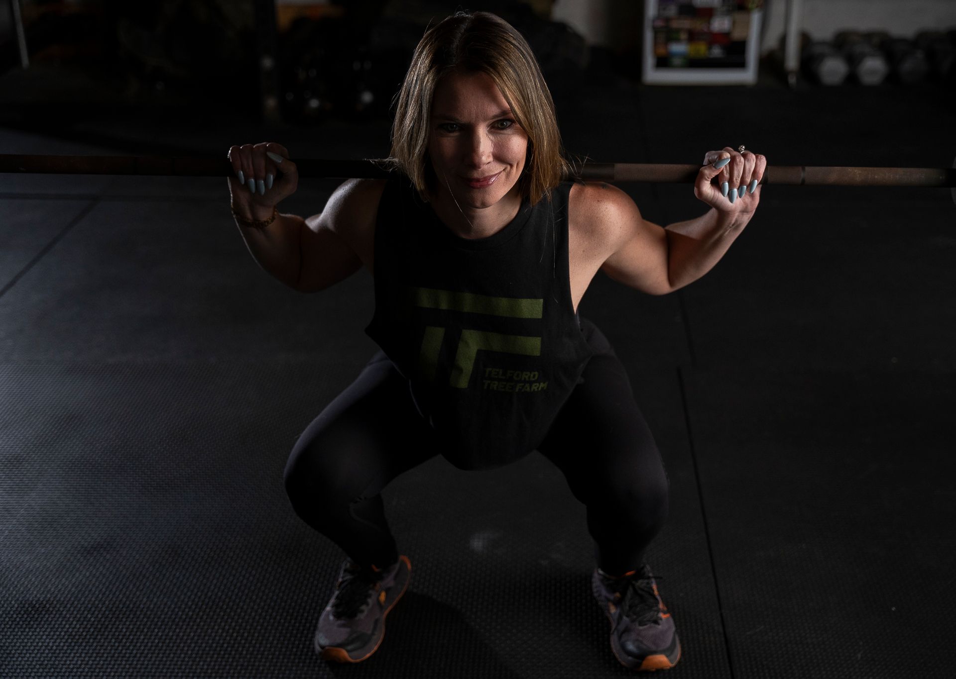 Woman squatting with a barbell in a dim gym, wearing black workout clothes and smiling slightly