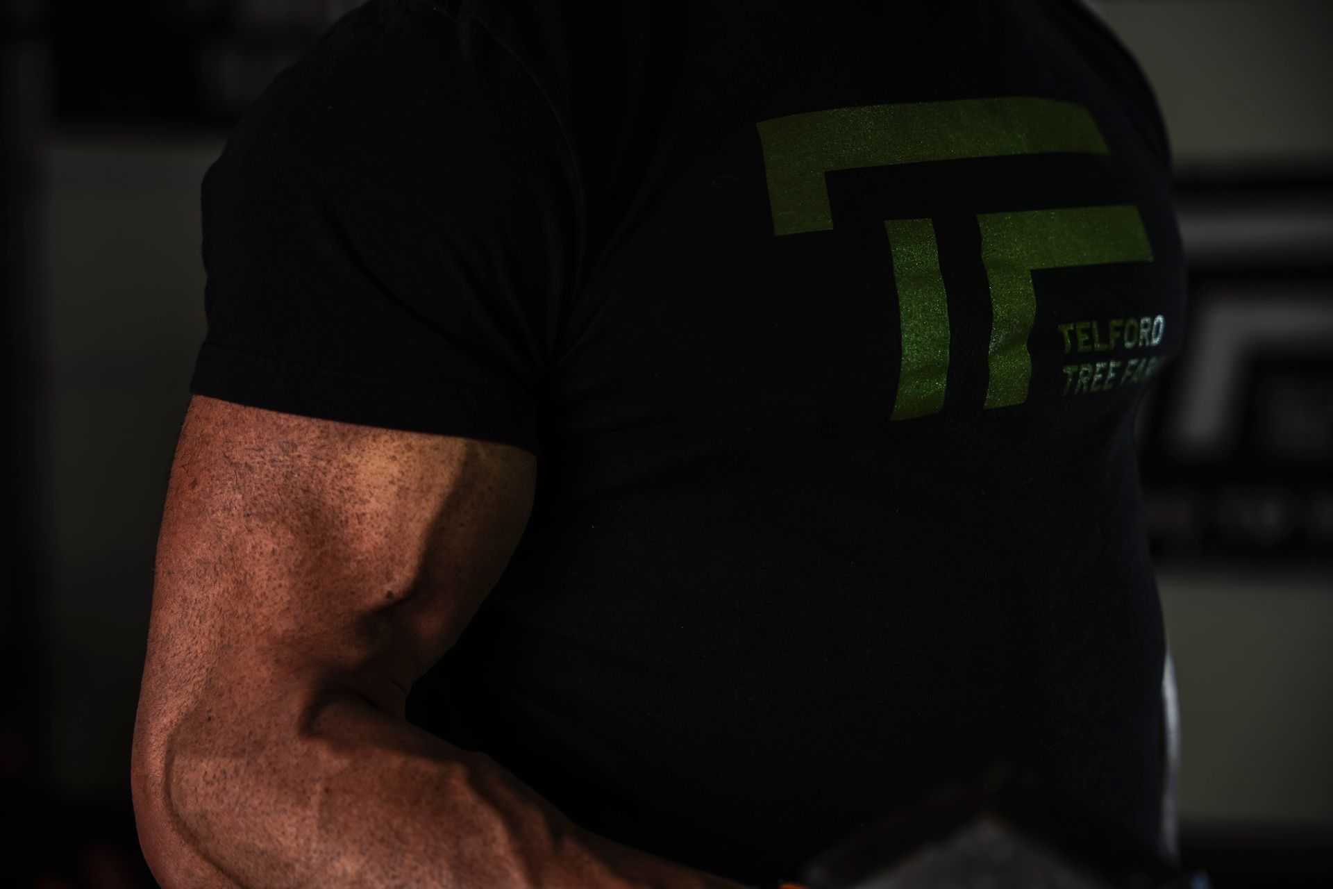 Close-up of a muscular arm in a black T-shirt with a green logo in low light