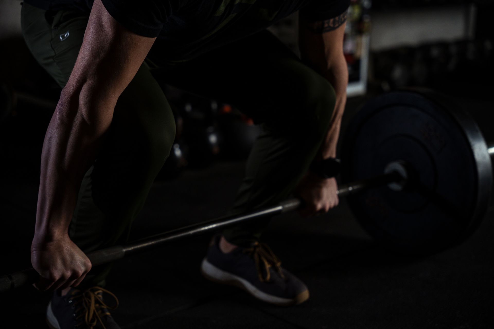Person lifting a barbell in a dark gym, close-up on hands and legs.