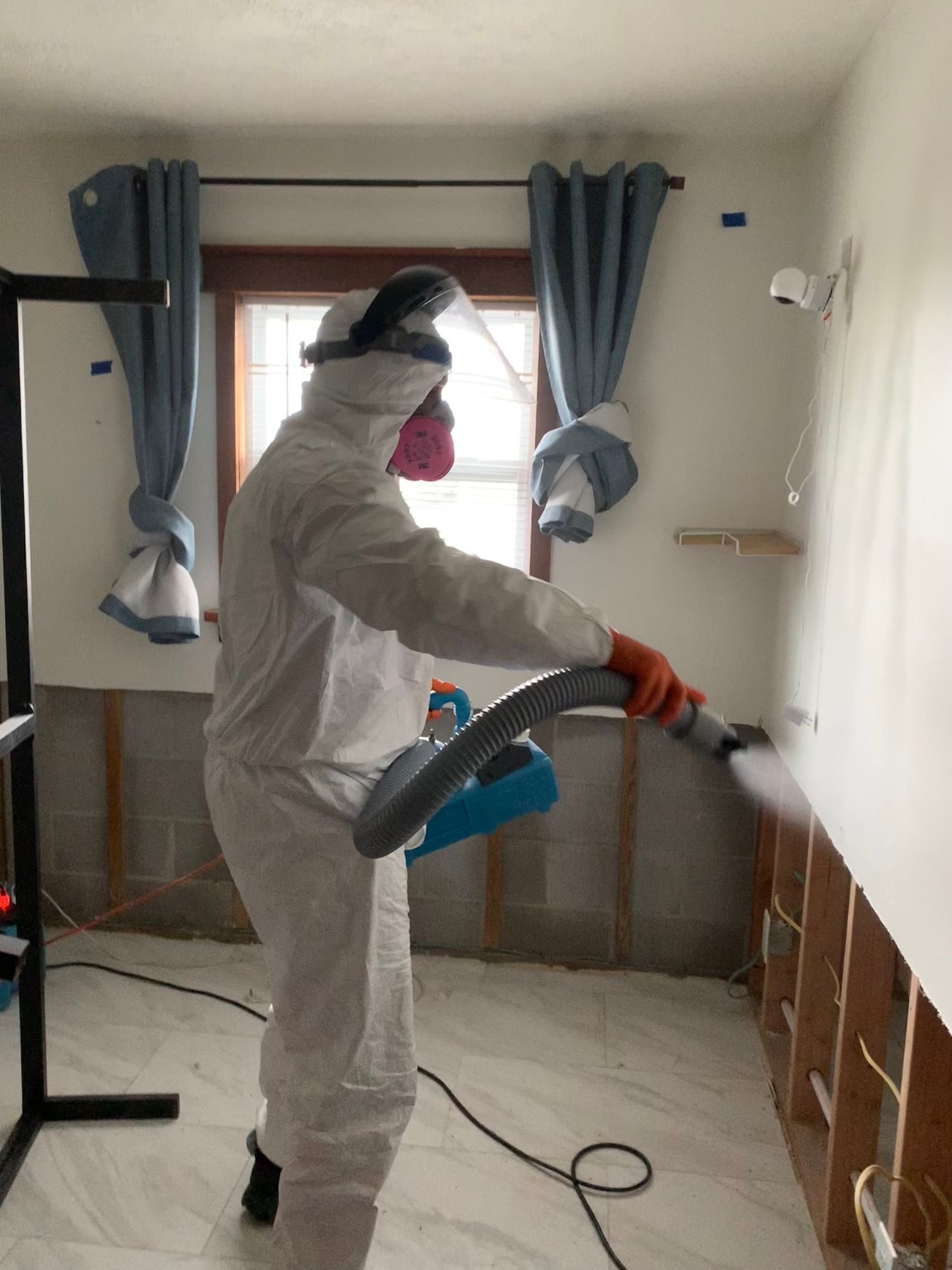 A person in a protective suit and respirator sprays a disinfectant solution onto the wall of a room with exposed studs.