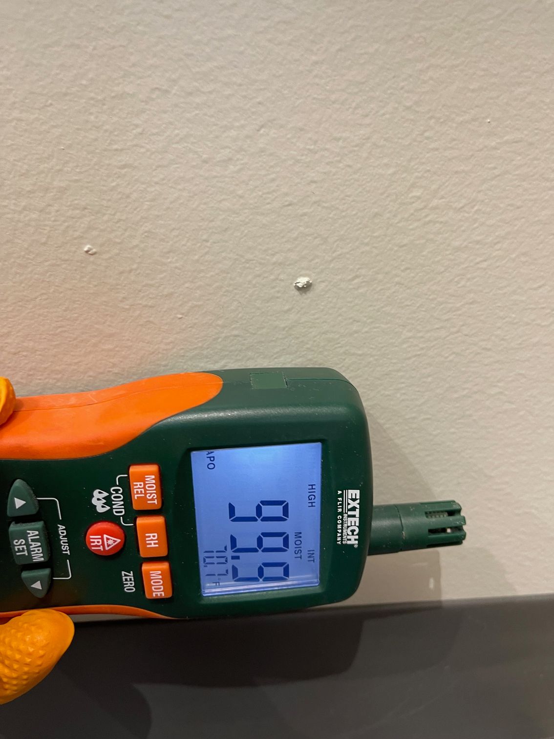 A green and orange digital moisture meter held against a wall, displaying a measurement of 99.9% on its screen.