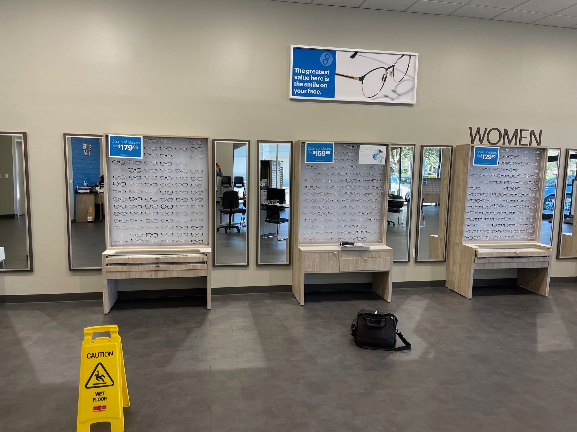 Eyewear store interior with wall-mounted displays of glasses, mirrors, and a yellow wet floor sign on a grey tile floor.