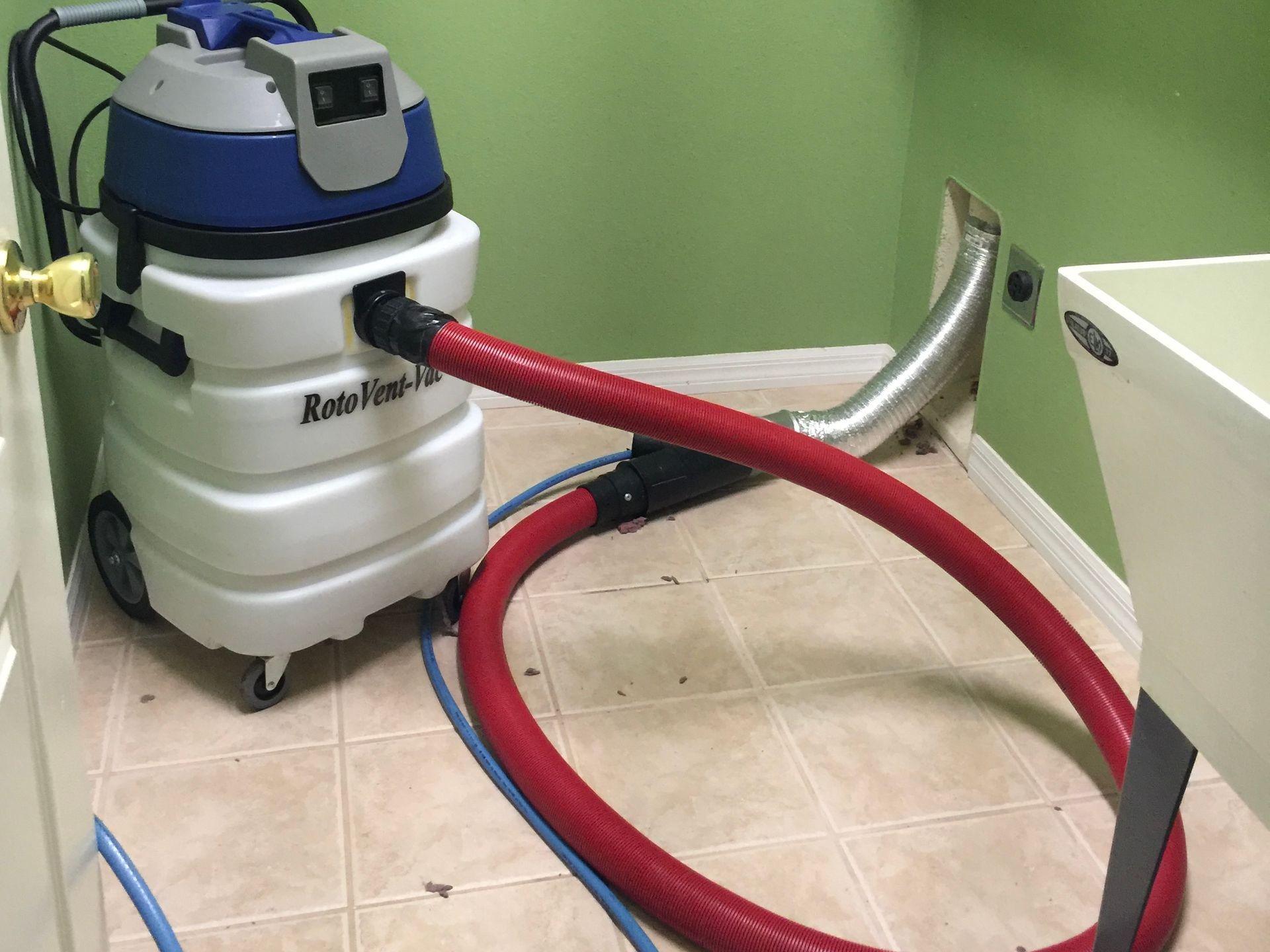 A wet-dry vacuum with a red hose connected to a silver vent pipe on the wall of a laundry room with a utility sink.