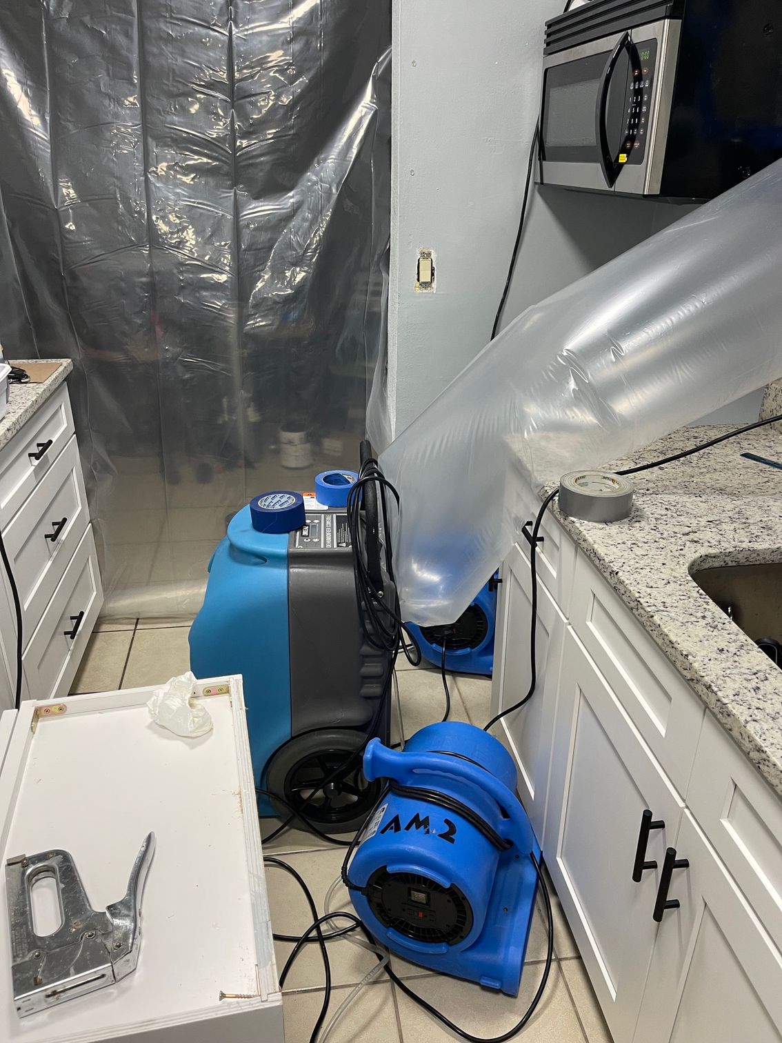 A kitchen undergoing water damage restoration with a blue dehumidifier, air movers, and plastic sheeting used for containment.