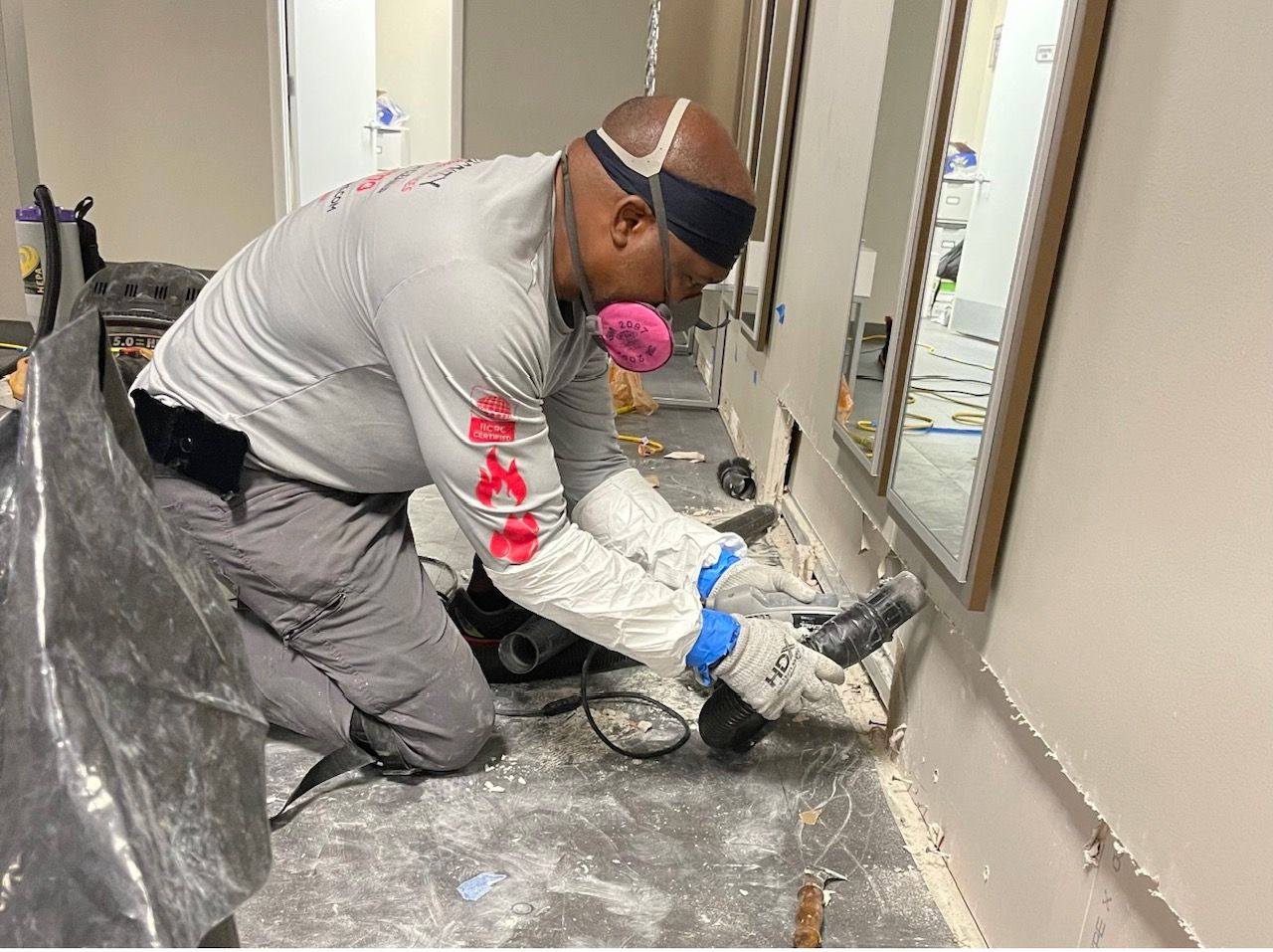 A worker wearing protective gear and a respirator uses a power tool to cut along a wall at floor level in a room.