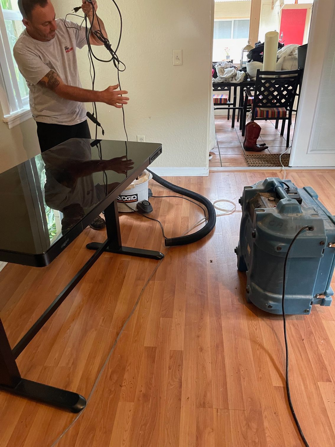 A person works in a room with a desk, hoses, and a large air-scrubbing machine on a wood floor during restoration work.