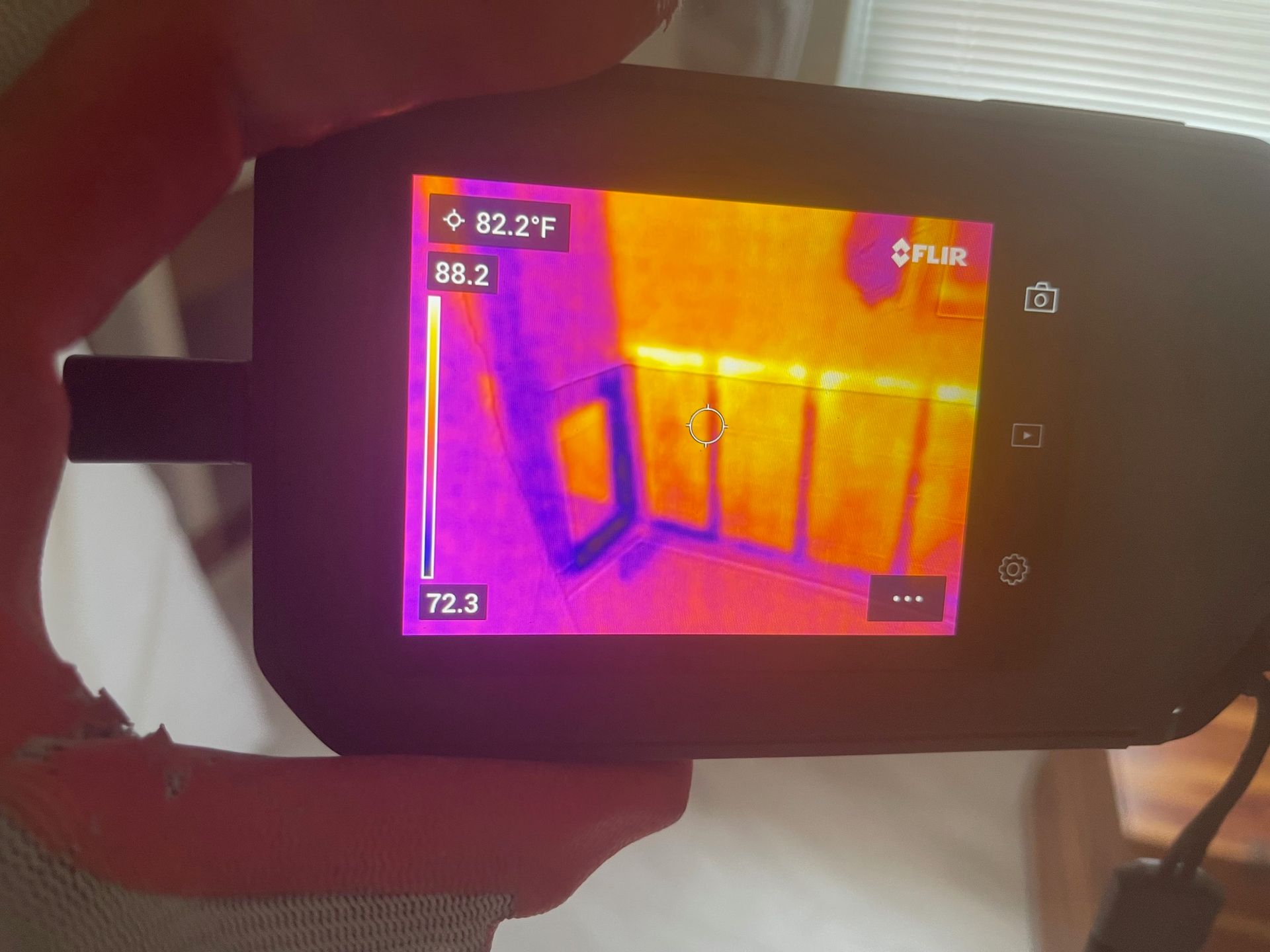 A hand holds a FLIR thermal camera displaying a heat map of a wall with visible studs, ranging from 72.3°F to 82.2°F.