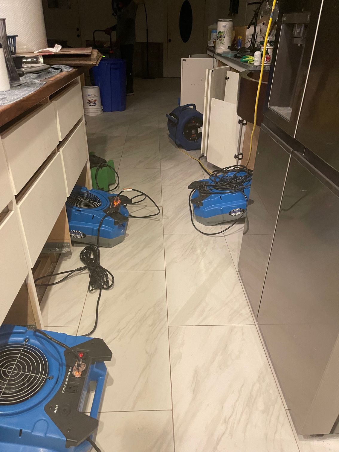 Several blue industrial air movers are positioned on a tiled kitchen floor during water damage restoration.