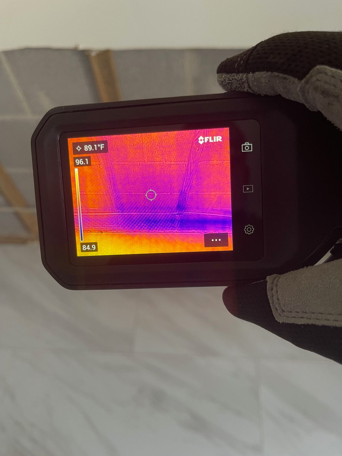 A gloved hand holds a thermal imaging camera displaying a heat map of a wall with vertical stud lines and a horizontal beam.