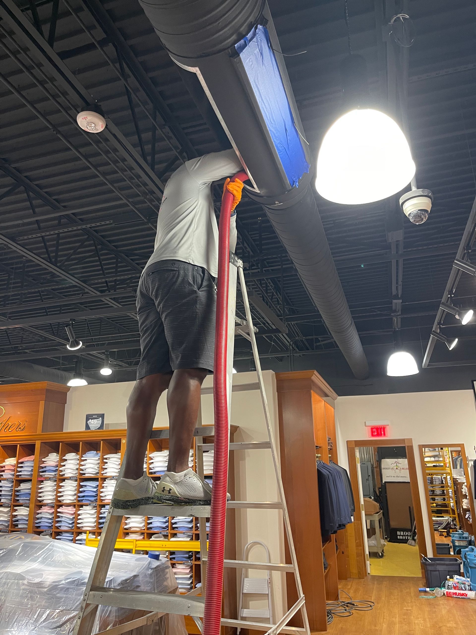 A person on a ladder reaches into an overhead HVAC duct in a clothing store while holding a red cable.