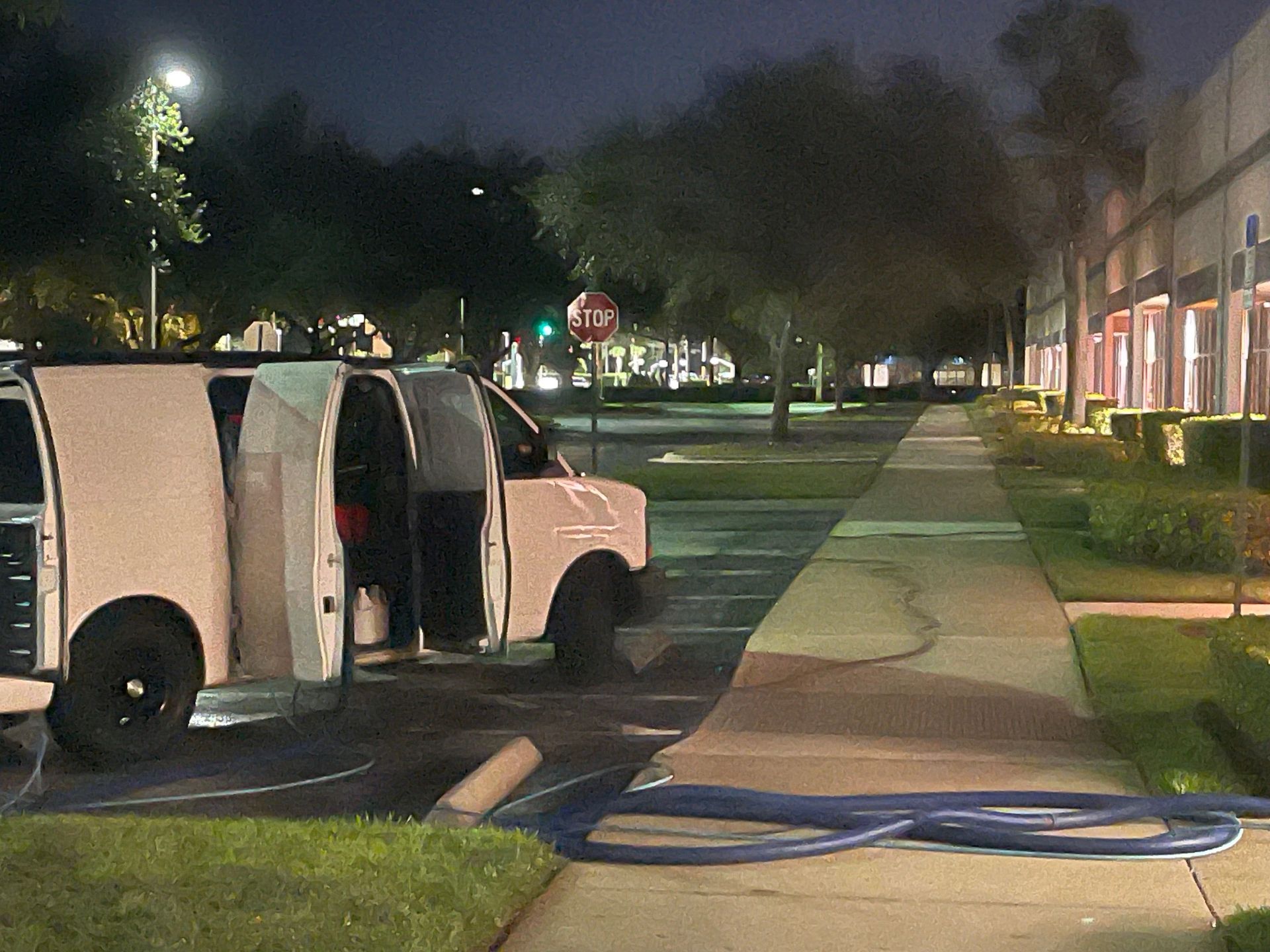 A white service van parked at night next to a sidewalk with a long blue hose stretched across the path.