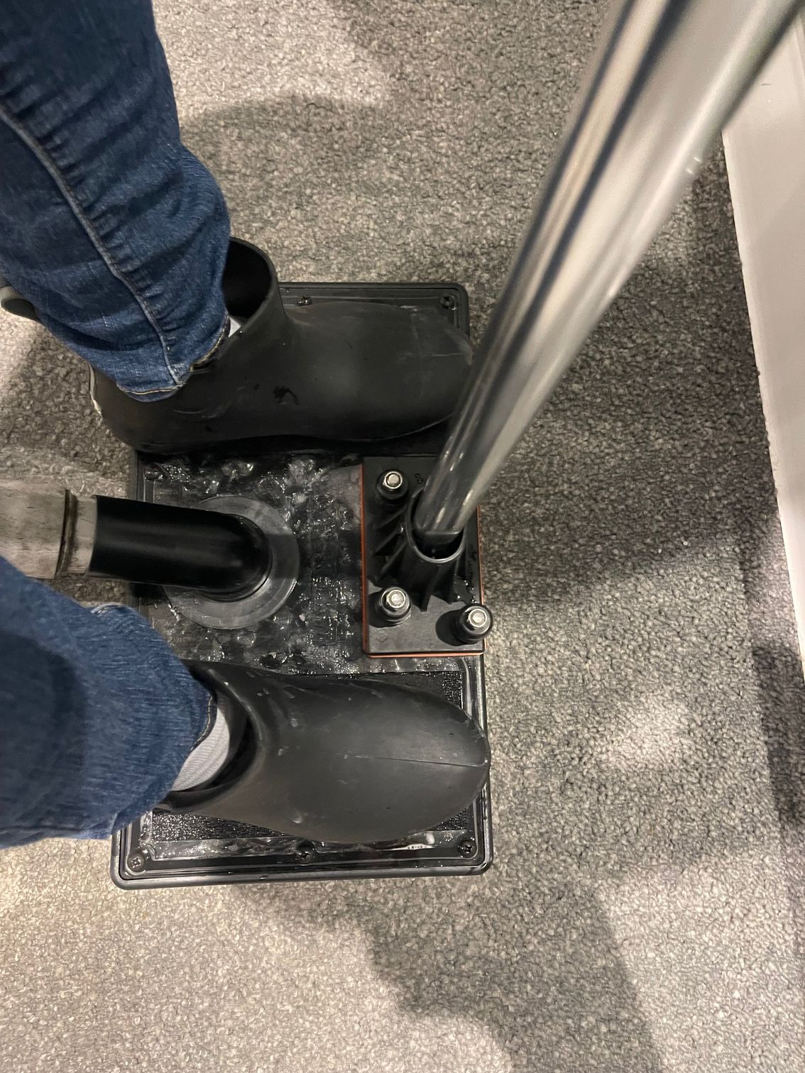 A person stands on a black, rectangular foot-operated pump device with a vertical handle on a grey carpeted floor.