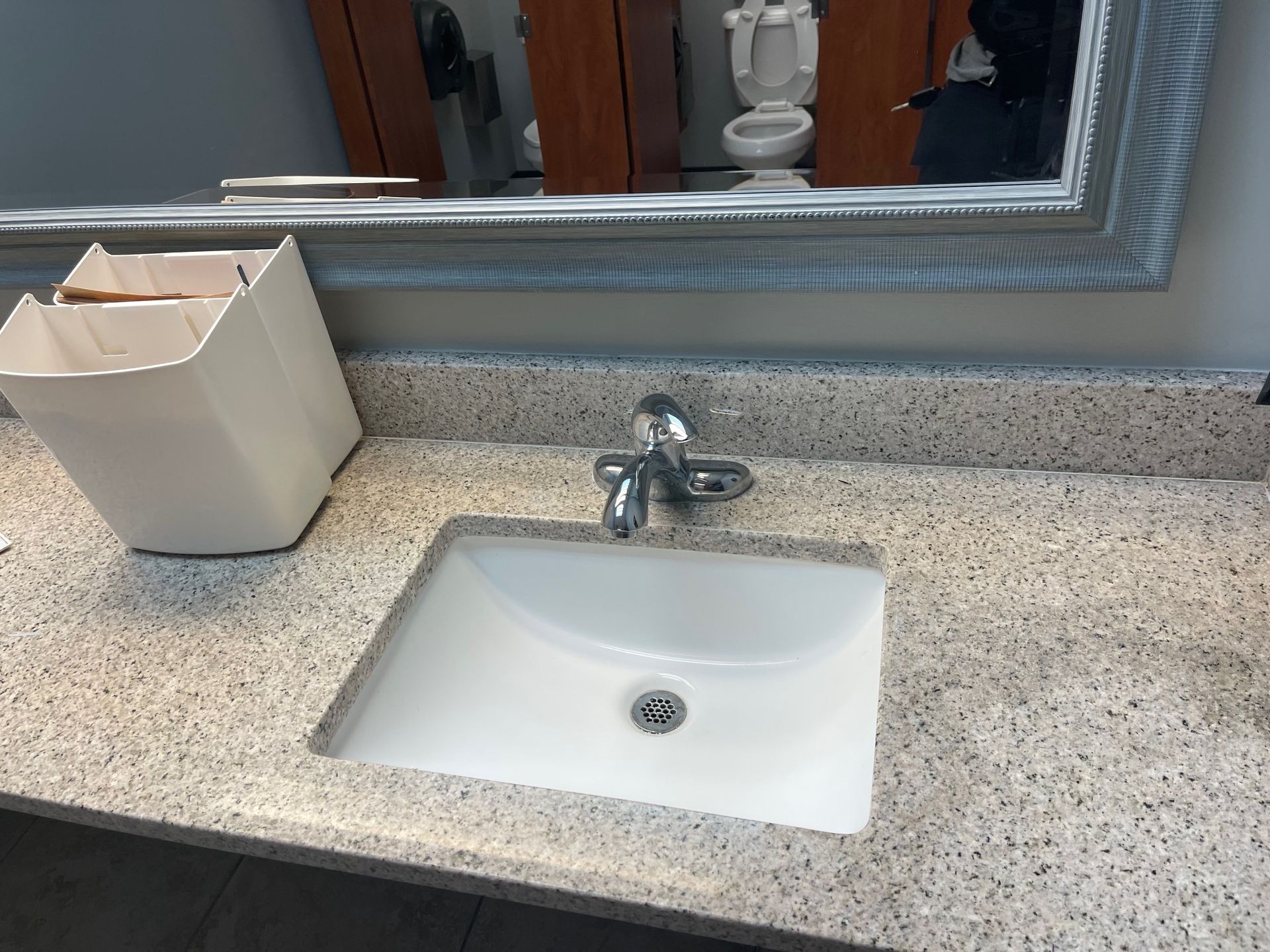 A white bathroom sink set in a speckled gray countertop with a matching wastebasket and a mirror reflecting a toilet.