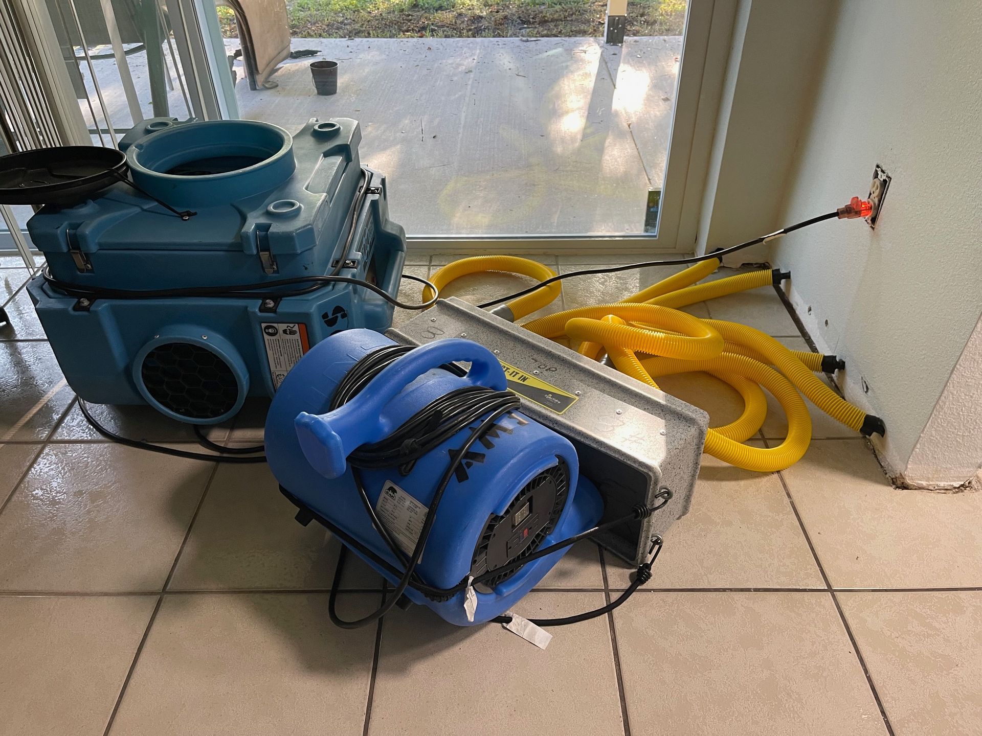 Blue industrial air movers and a dehumidifier are set up on a tiled floor near a sliding door, plugged into a wall outlet.