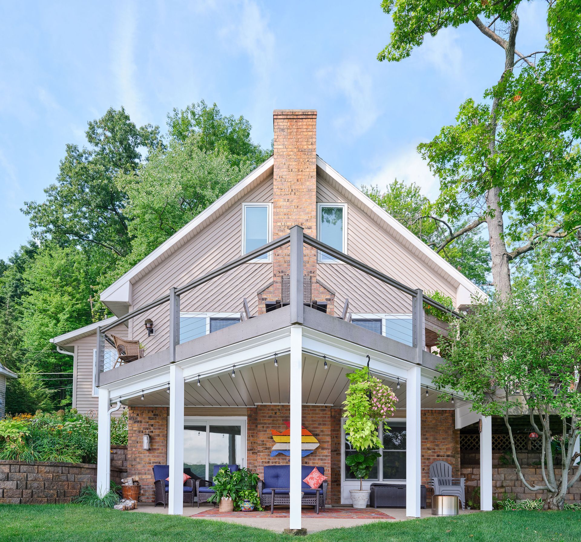 Two-story beige house with a brick chimney, white porch, and deck surrounded by green trees and lawn