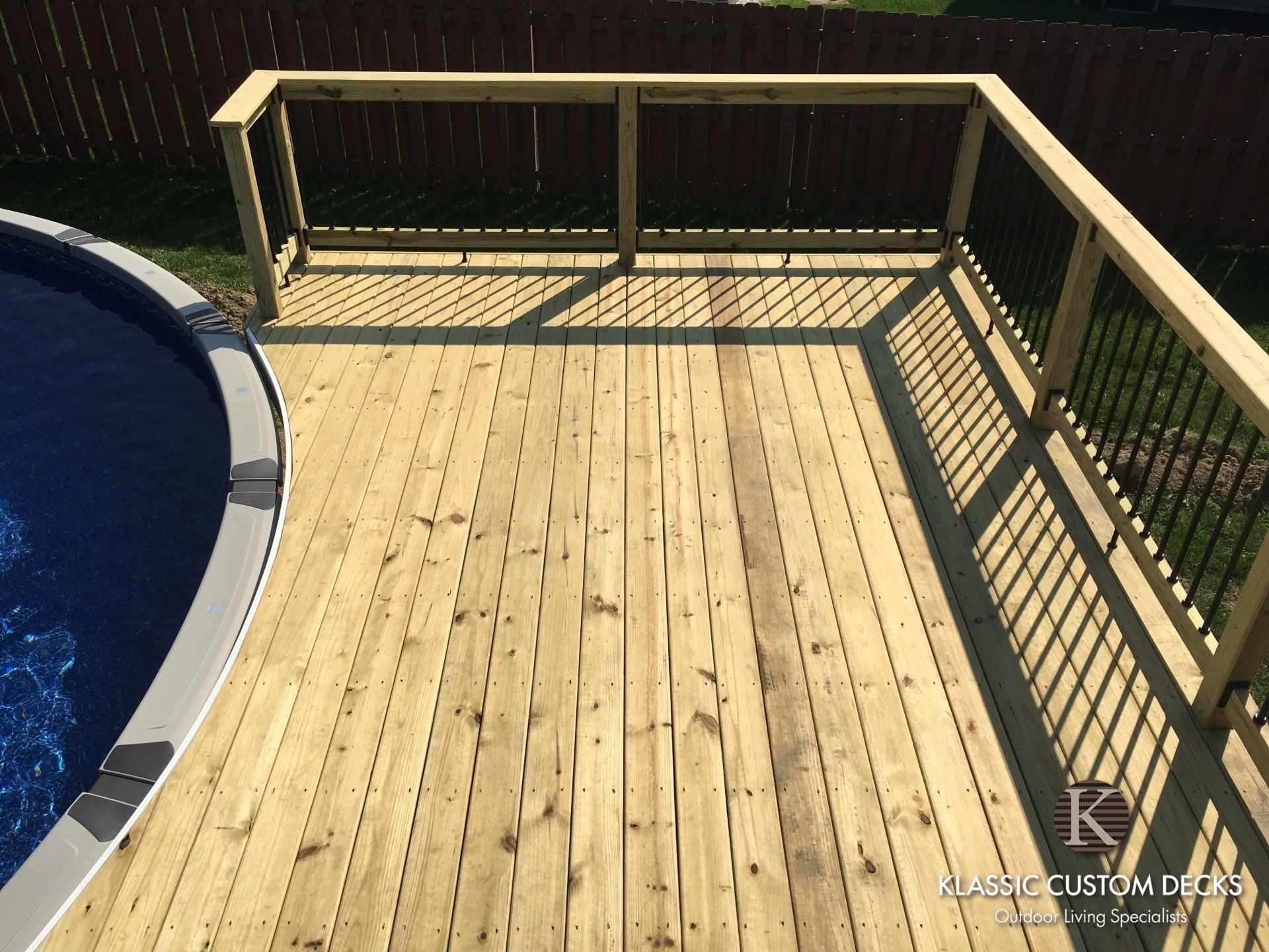 Wooden pool deck with railing beside a blue swimming pool in bright sunlight