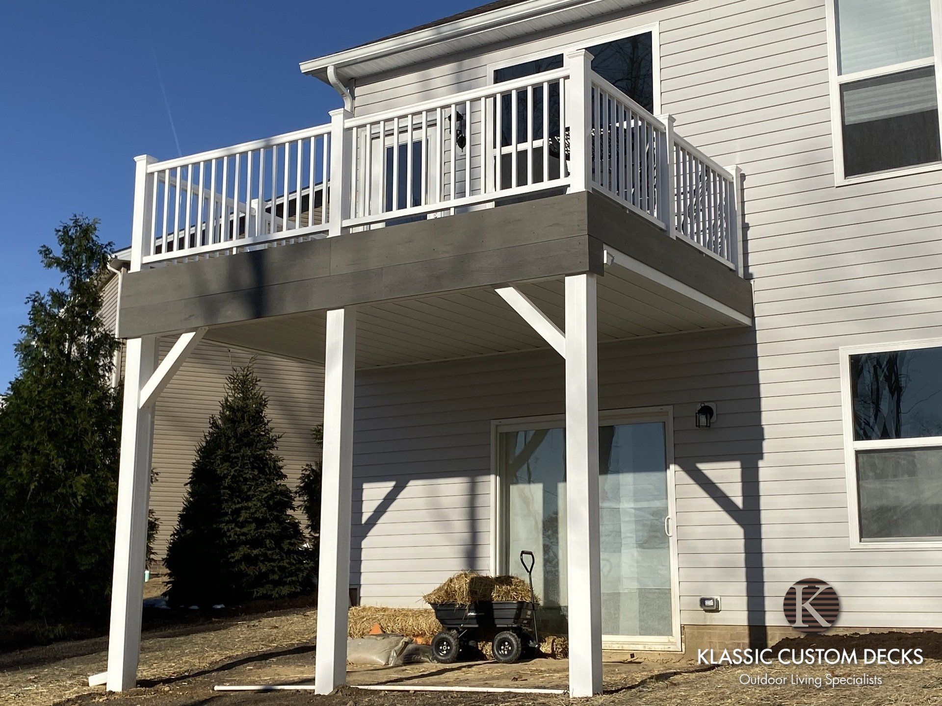 Two-story beige house with a white-railed deck on stilts and a small patio below