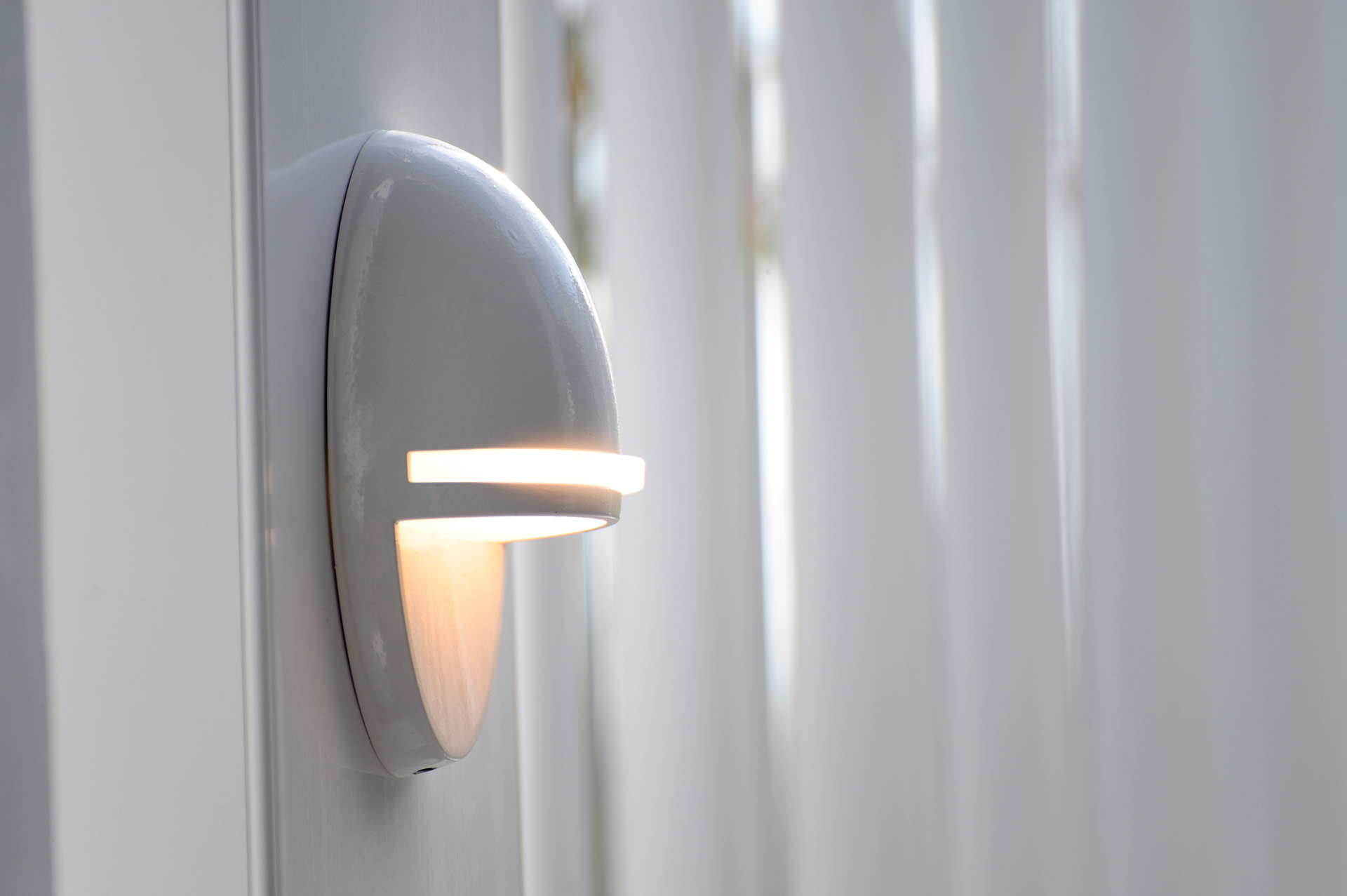 Wall-mounted gray light fixture glowing beside white vertical blinds