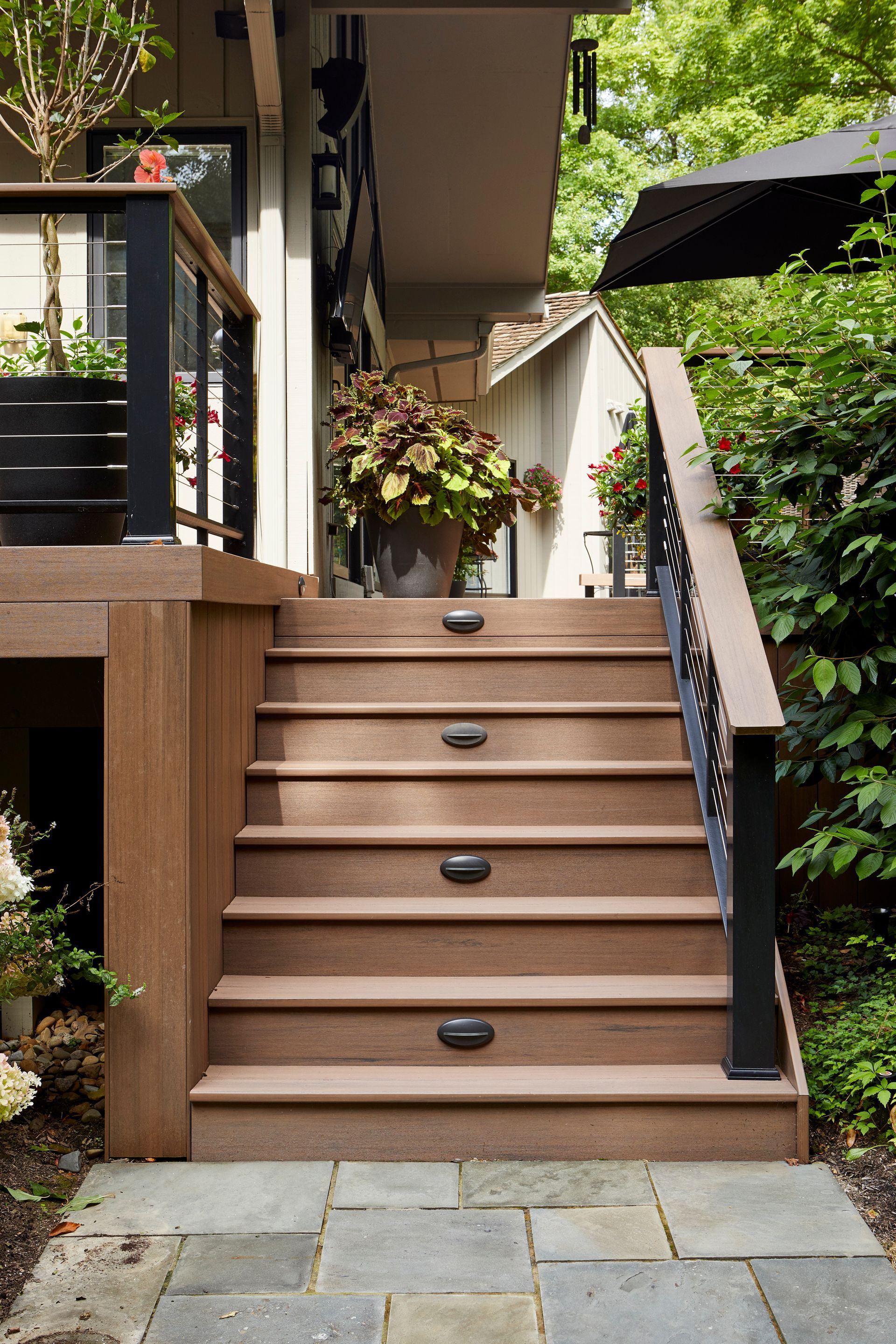 Wooden outdoor stairs with black railings leading up to a house, surrounded by greenery and potted flowers.