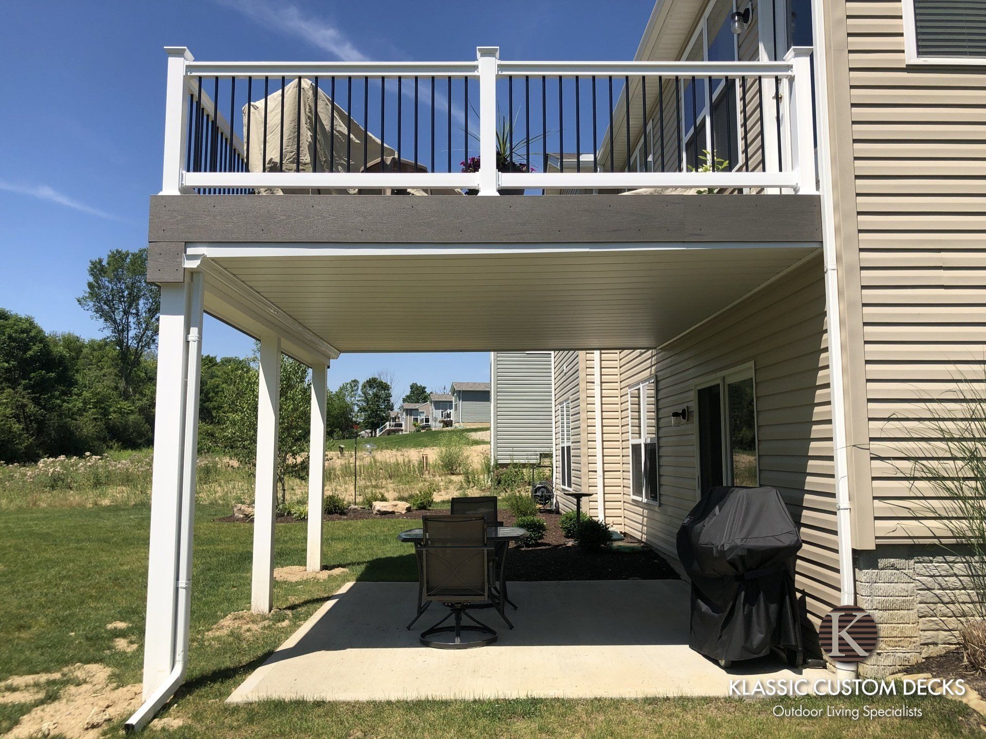 Back patio under a raised deck with white railings, beige siding, grill, and patio furniture