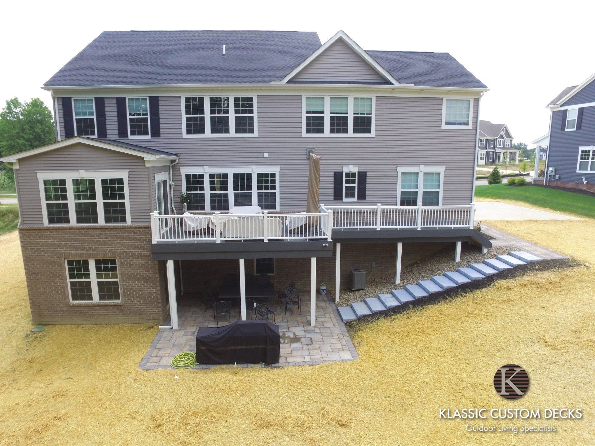 Rear view of a gray two-story house with white trim, deck, and patio over a bare yellow yard
