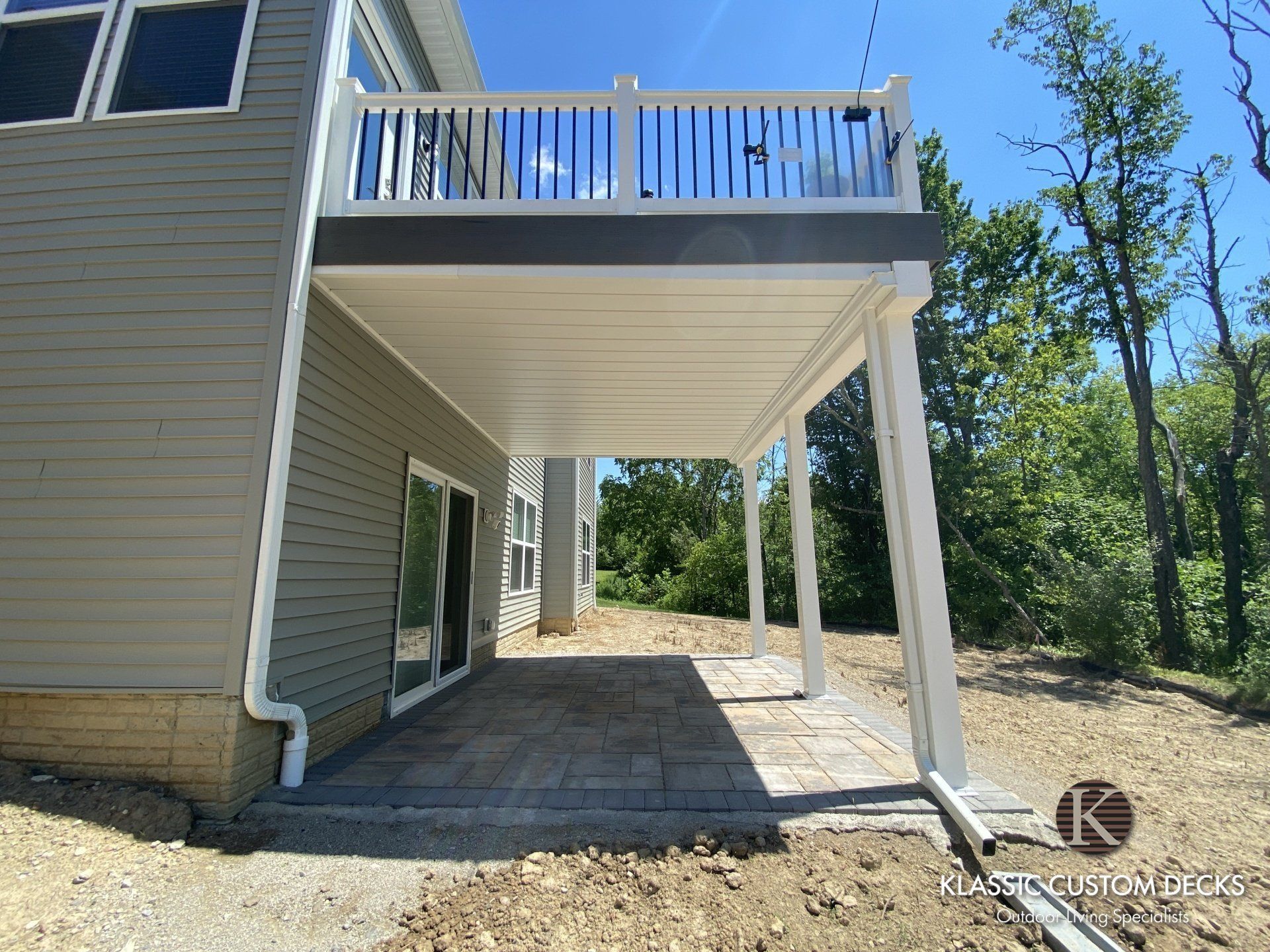 Back deck with white railing and support posts beside a beige house, overlooking a wooded area